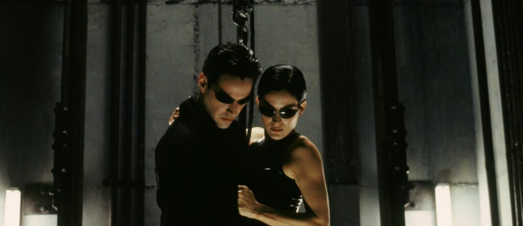 Affiche du film Matrix des sœurs Wachowski. Quatre personnages en vêtement noirs latex et lunettes de soleil, dont un à l’avant tient une arme.